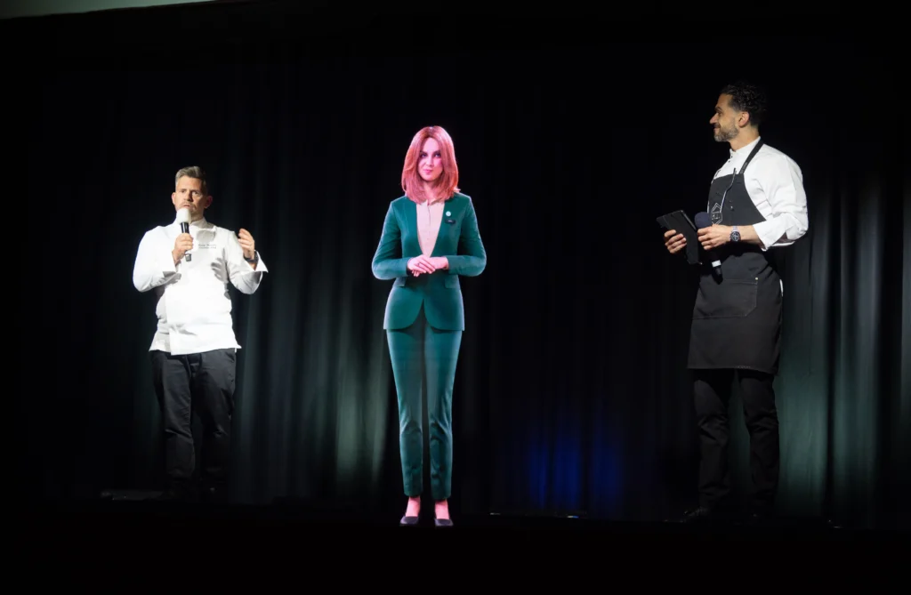 AI hologram Anna stands between two chefs presenting on stage at the Pullman London dinner