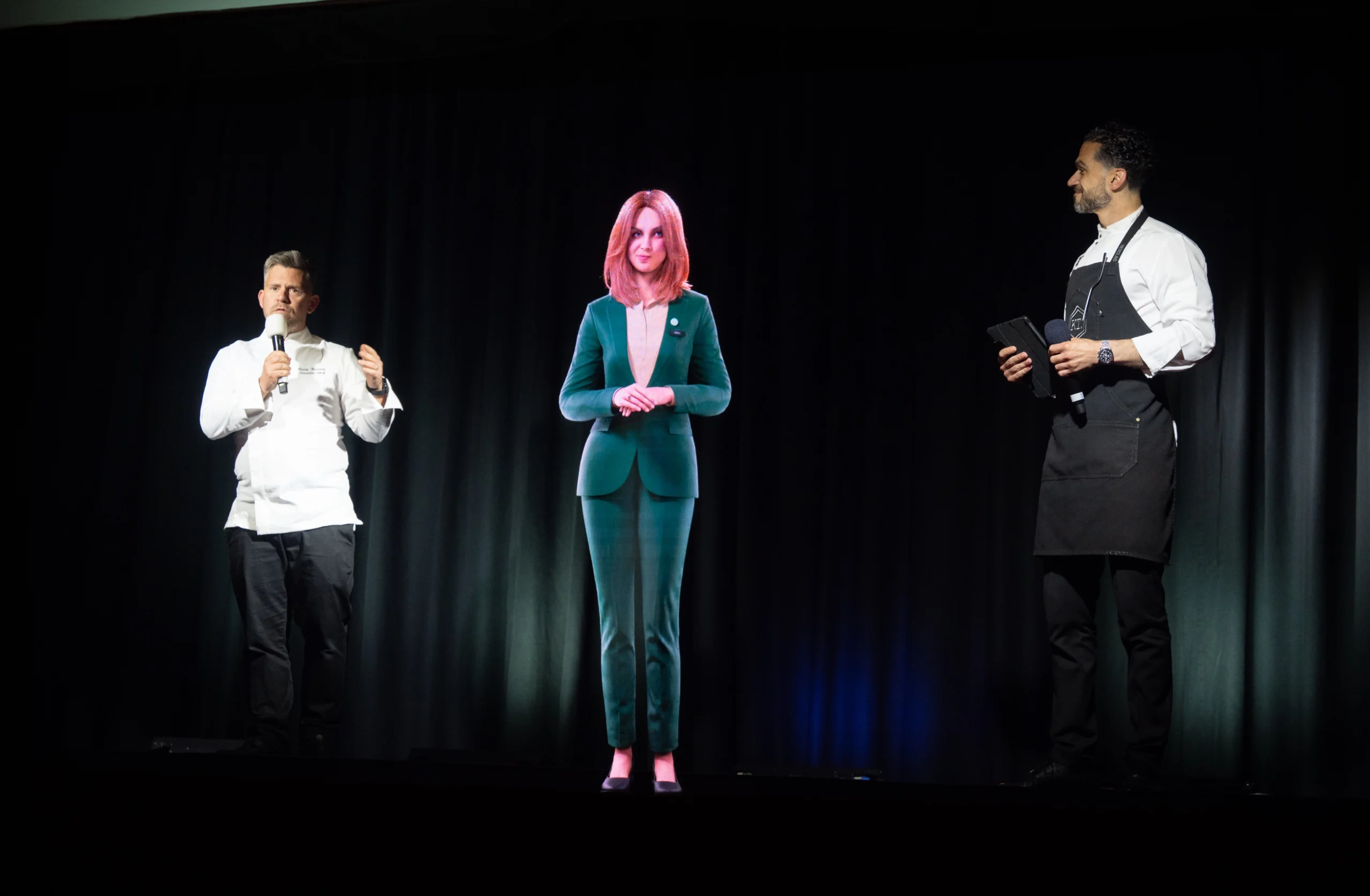 AI hologram Anna stands between two chefs presenting on stage at the Pullman London dinner