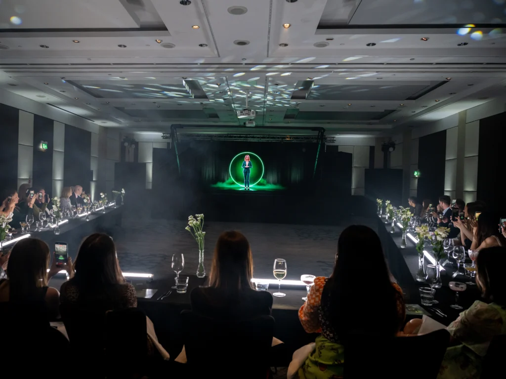 Guests watch Anna, RAVATAR's AI hologram, glowing in a green ring on stage at Pullman London