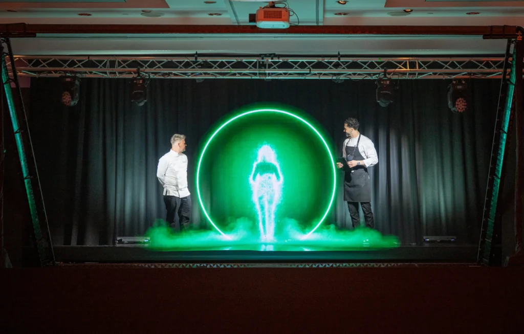 Two chefs stand beside AI hologram Anna as she forms in a glowing green ring of mist on stage at Pullman London