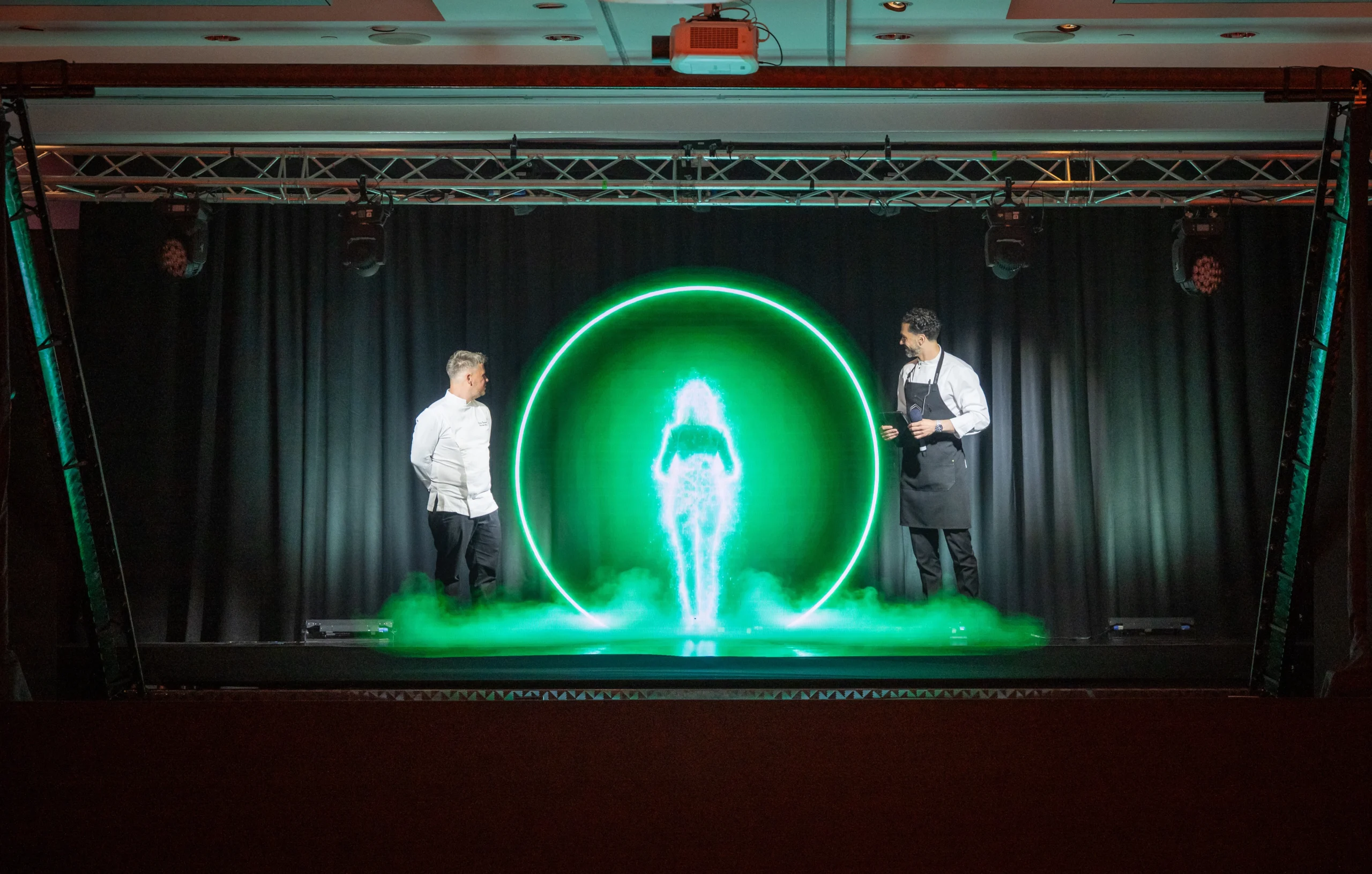 Two chefs stand beside AI hologram Anna as she forms in a glowing green ring of mist on stage at Pullman London